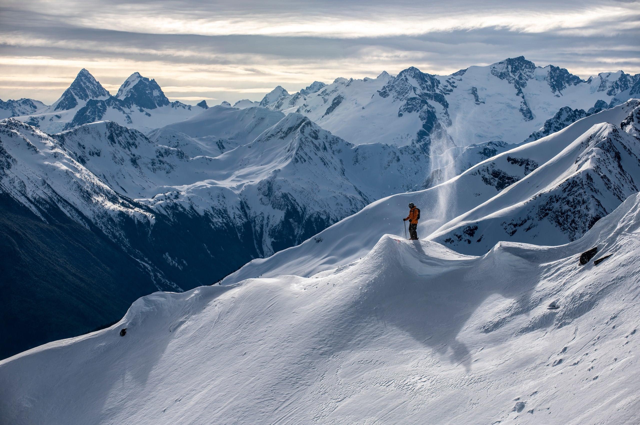 a skier in an orange jacket resting on a flat surface looking over the mountain