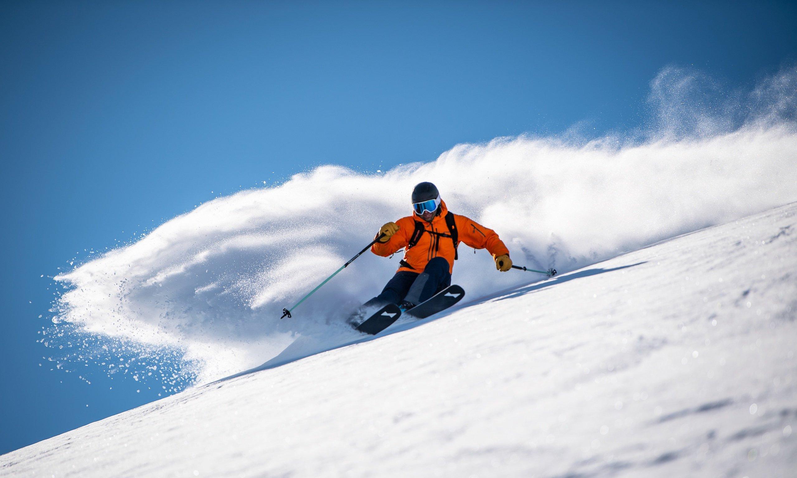 A skier in an orange jacket and helmet descends a snowy slope, kicking up a spray of snow under a clear blue sky.