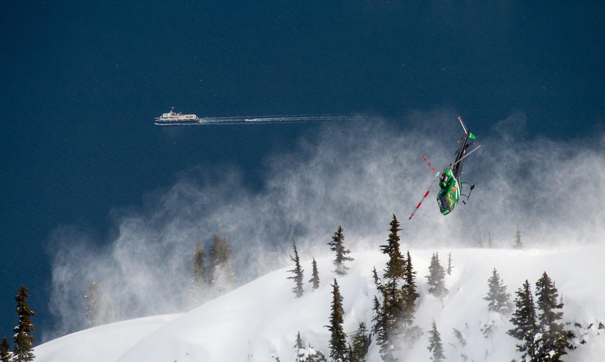 A helicopter flying over a mountain while a boat sails in the water next to the mountain