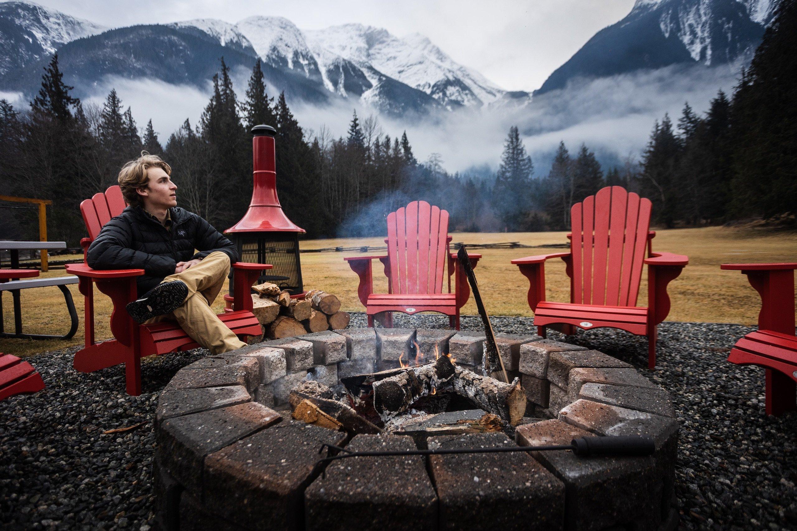 A man on a red chair sitting near the fire with a misty mountain range in the background