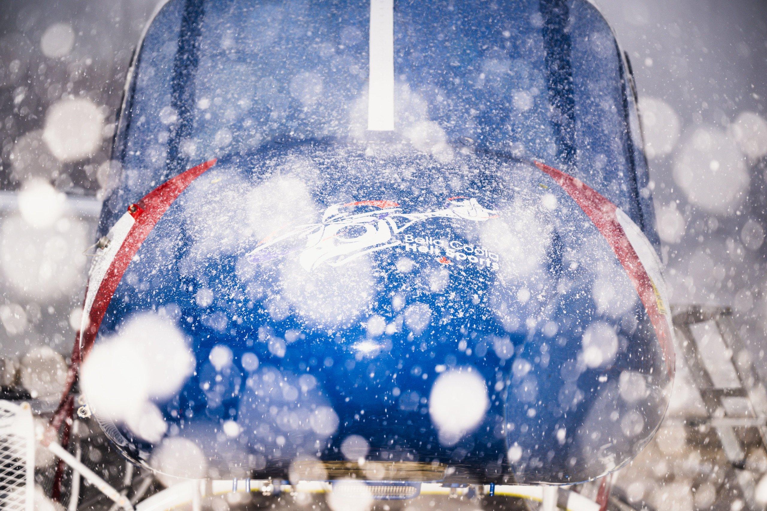 Snow falling down in front of a blue helicopter with the Bella Coola Heli Sports logo