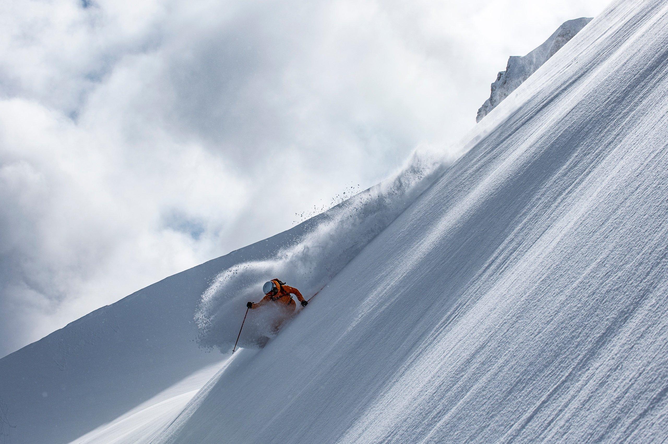 a person in an orange snowsuit skiing down a mountain leaving a plume of snow in their wake