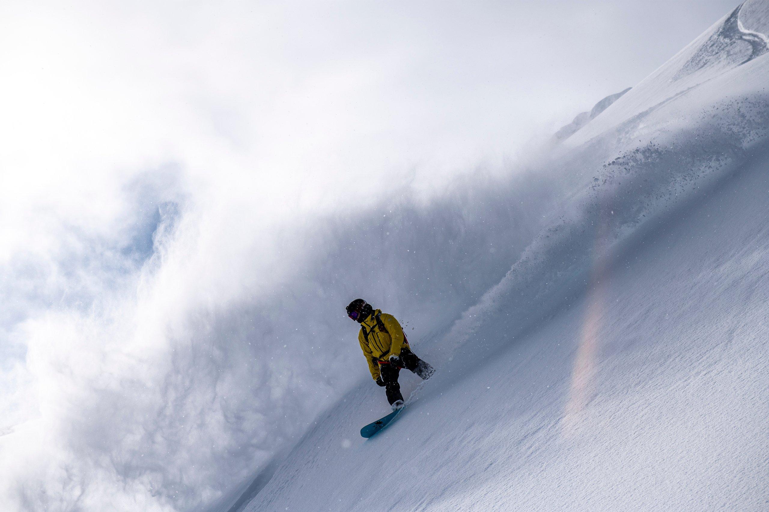 a ski guide in a yellow jacket riding a snowboard down a mountain in powder snow