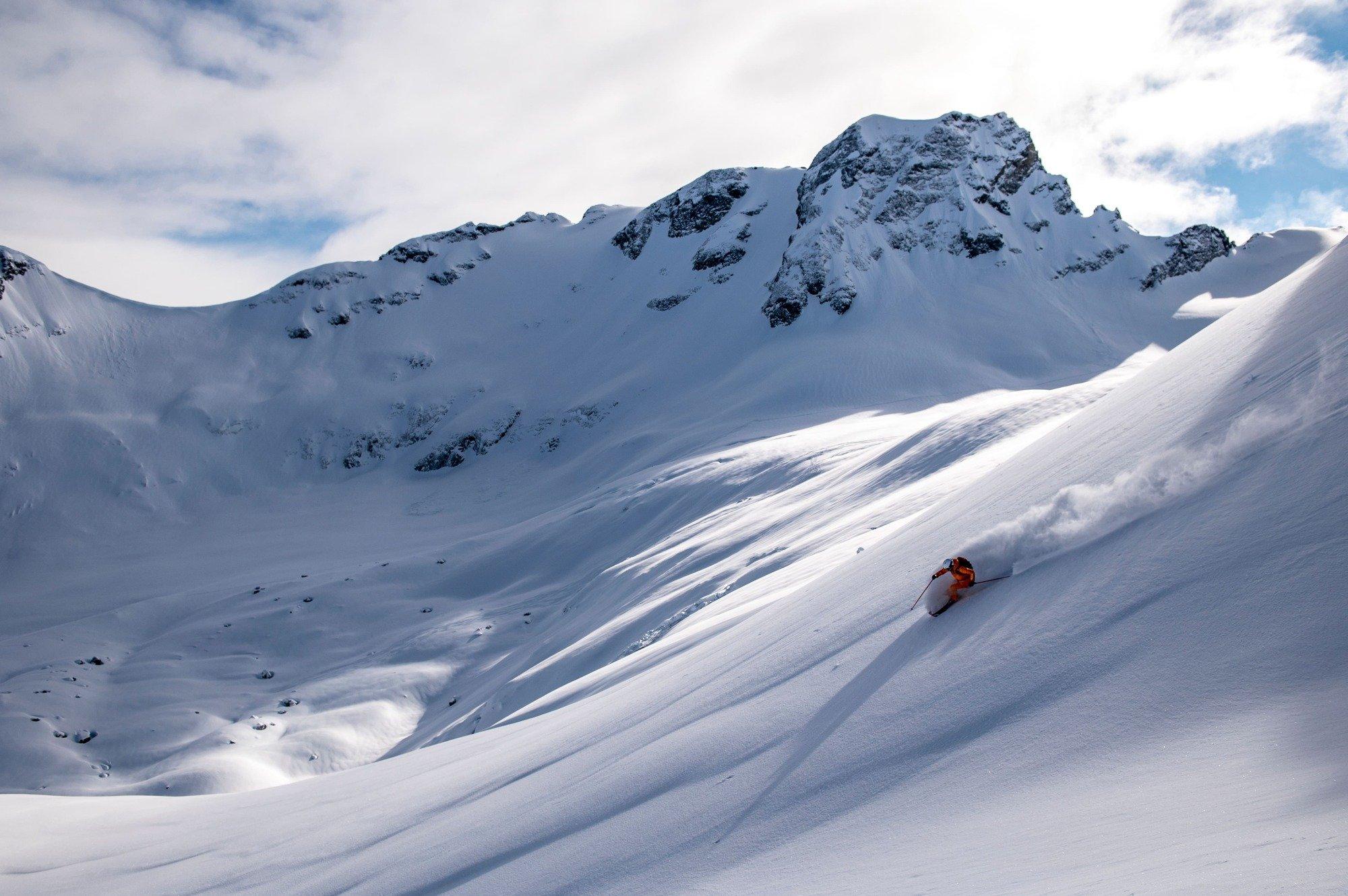 A skier in an orange snowsuit descending a mountain leaving a plume of snow trailing behind them