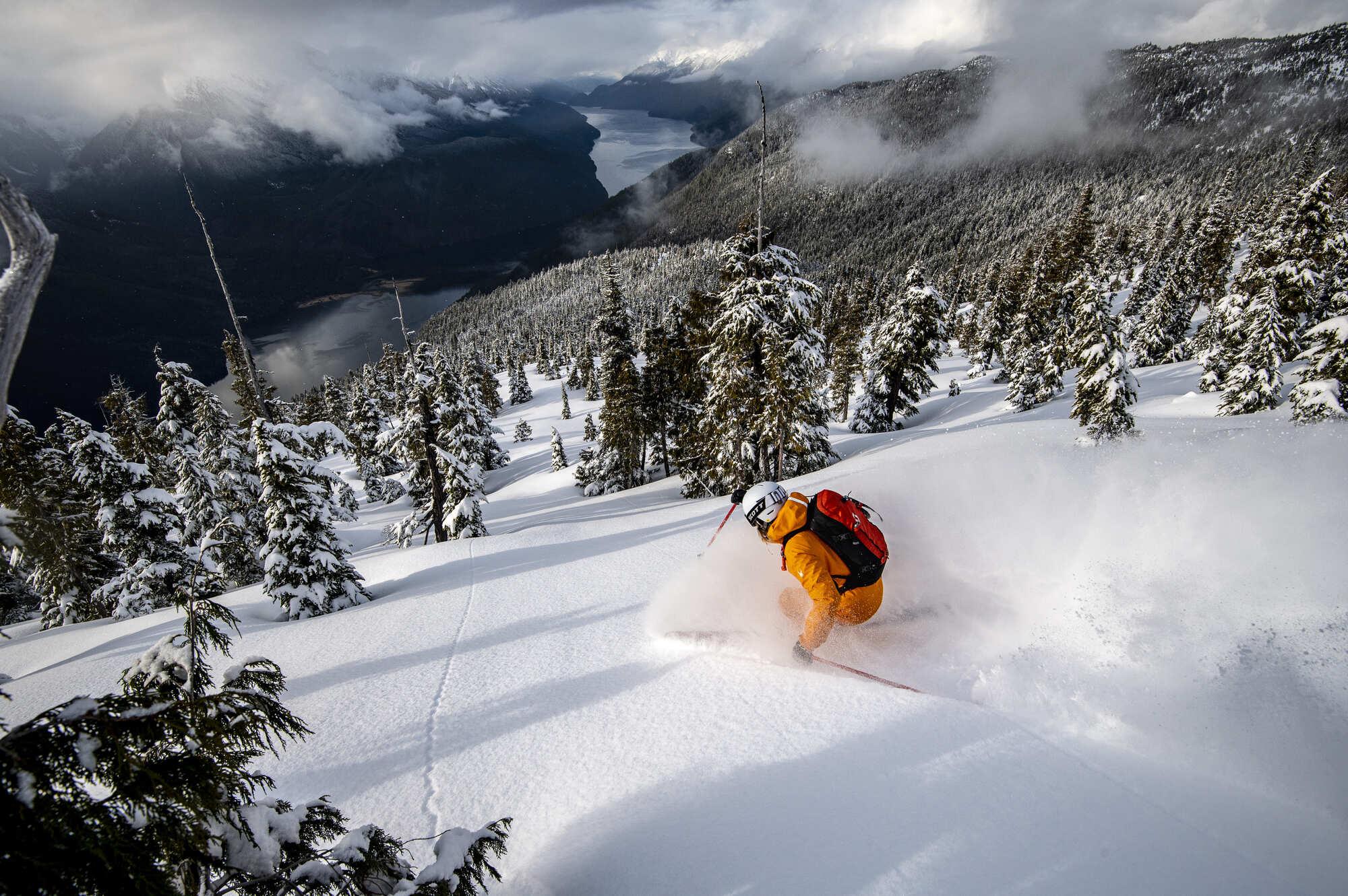 Skier in an orange snowsuit and red backpack descending a tree filled mountain