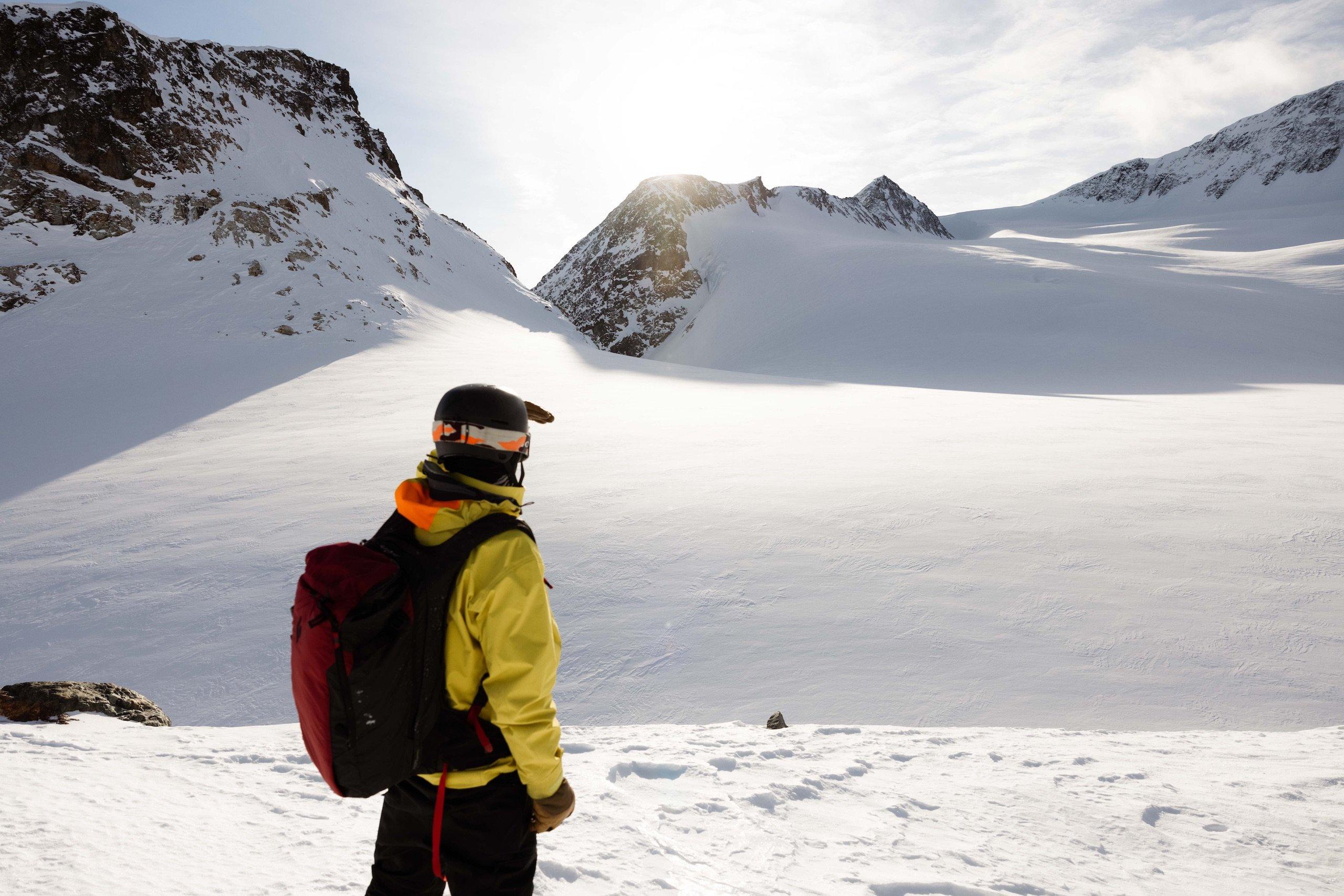 A skier in a yellow jacket and red backpack resting and looking at the mountain