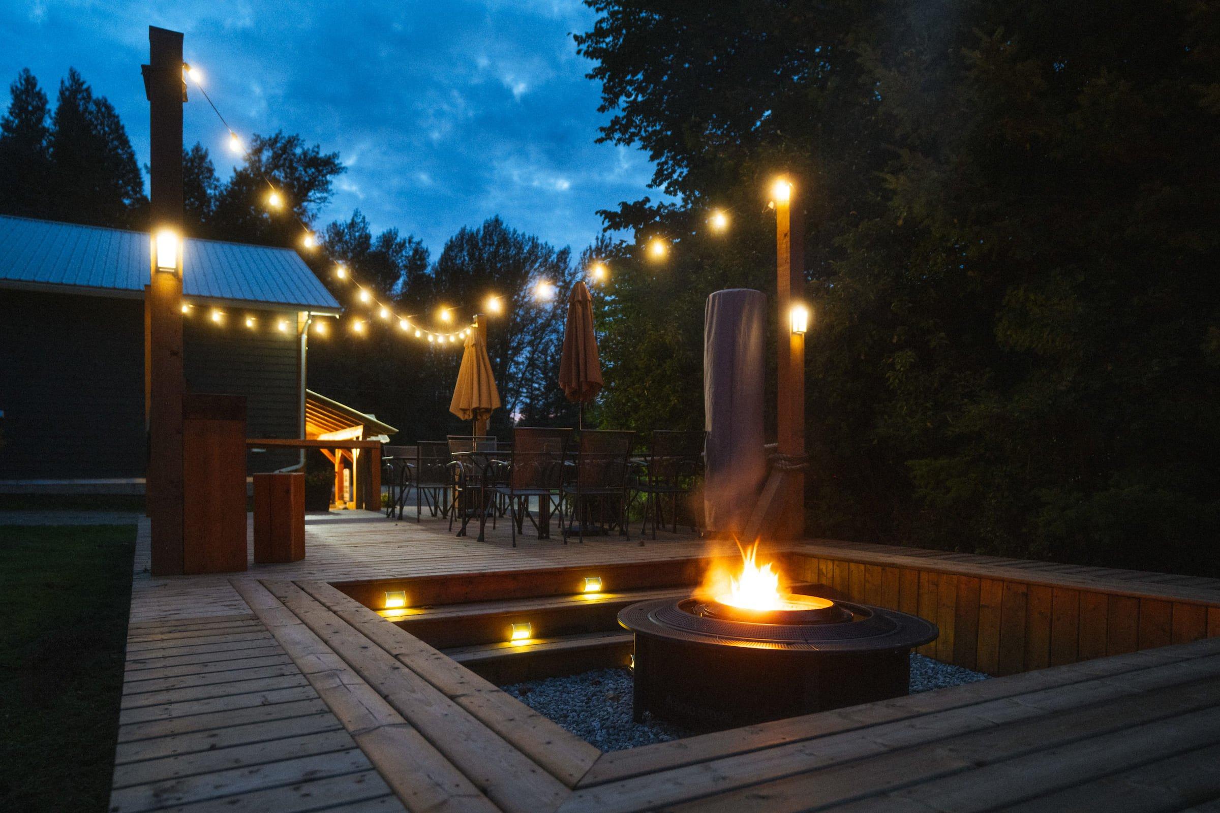 A large outdoor patio with overhead string lights and a propane fireplace surrounded by build in benches